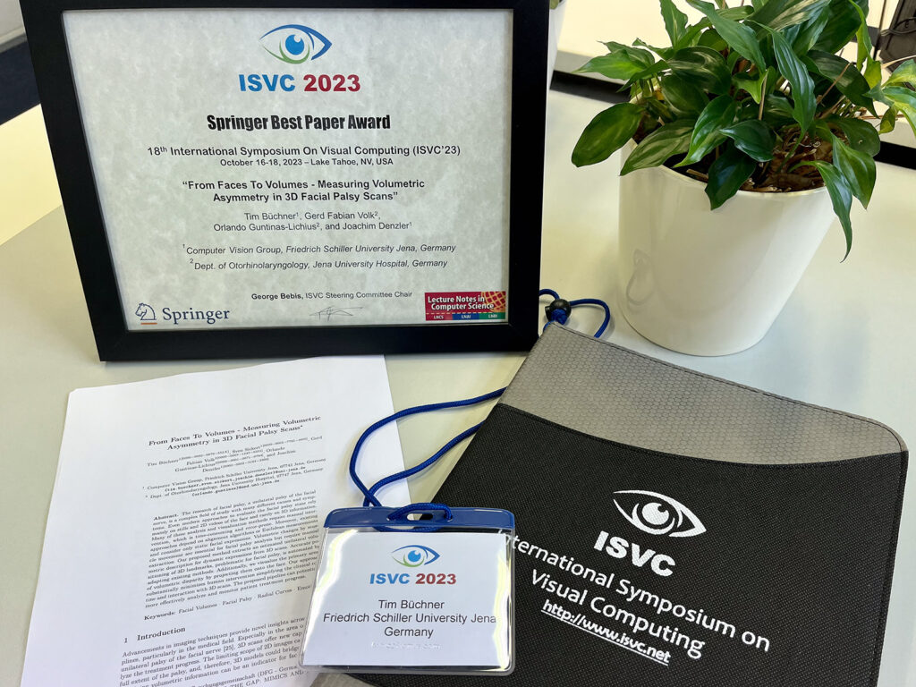 The image depicts a collection of items from the ISVC 2023 conference. In the upper left corner there is a framed version of the best paper award certificate. Next to it on the right is a plant as a decorative element. In the lower part of the image there is a print of the paper on the left and a writing case, which has ISVC inscriptions on it. In between those two items is the name badge of Tim Büchner, who attended the conference.